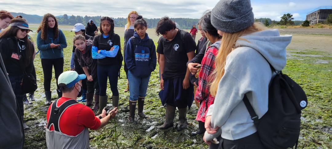 High school students stand on a beach looking at a man in a red shirt holding an organism.