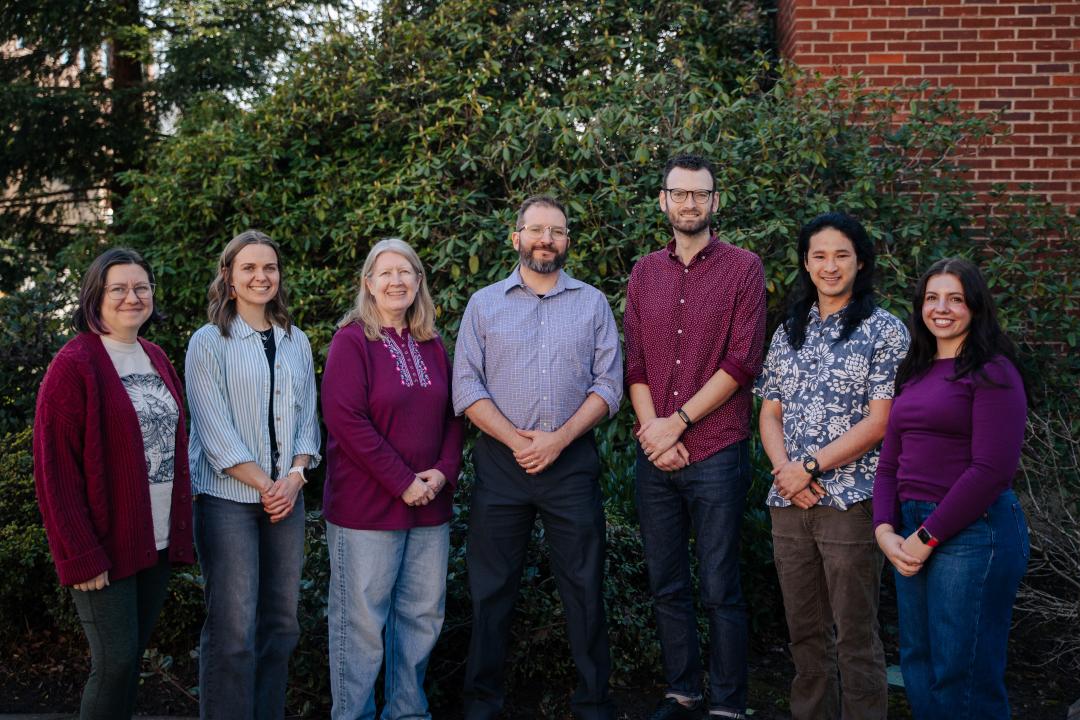 Seven members of the Sharpton Lab stand shoulder-to-shoulder outside in front of a large, green bush, each wearing matching maroon and blue shirts.