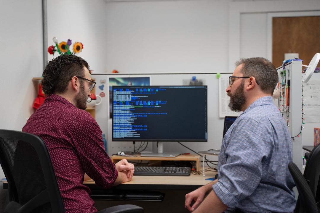 Two men sit at a desk with a monitor covered with code, discussing the information.