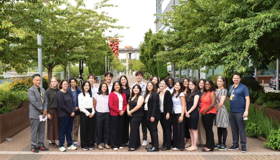 On a sunny day, a large group of people in business-casual attire stand and smile at the camera on a brick campus road lined with trees.