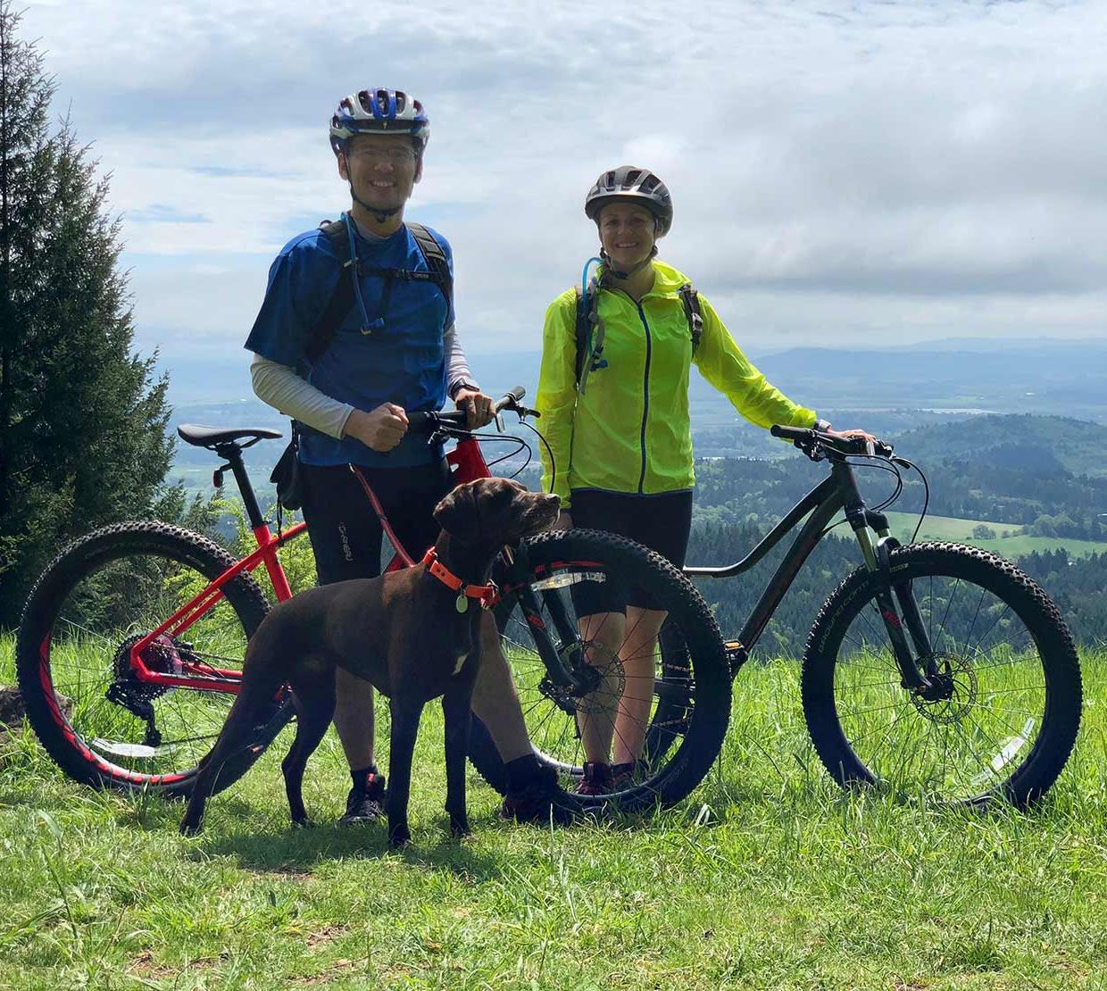 Courtney Rae Armour with husband and dog riding mountain bikes atop grassy hill