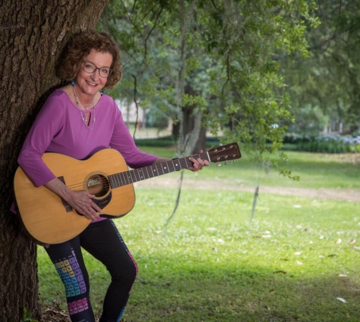 A woman in a purple shirt leaning against a tree and holding a guitar