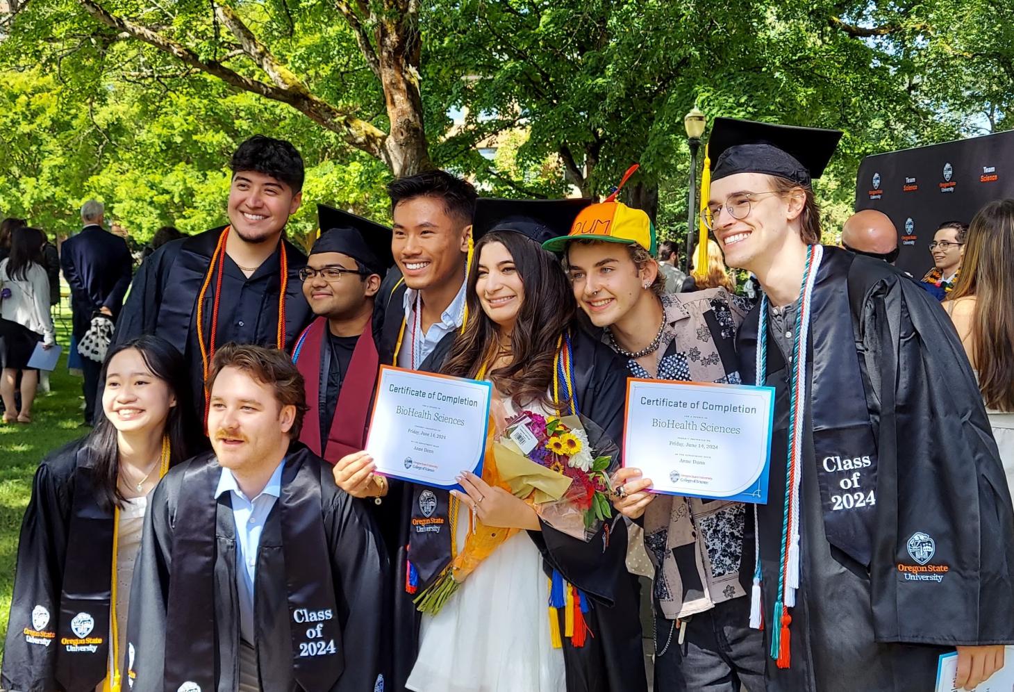 Group of students in graduation caps and gowns posing outside with certificates.