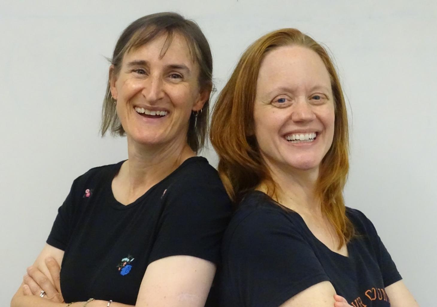 Linda Bruslind and Allison Evans, wearing black t-shirts, stand smiling back to back with crossed arms.