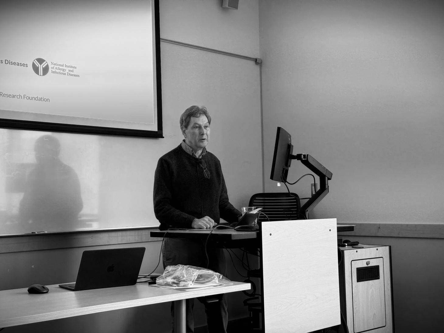 Scott Landfear, lecturing in a classroom with a projector screen behind him.