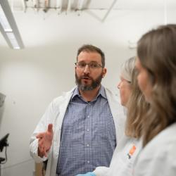 A bearded man wearing glasses and a button-up shirt beneath a lab coat speaks to two other scientists wearing lab coats toward the camera.