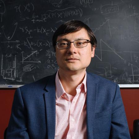 Jeff Gore, professor of physics at MIT, seated in front of a chalkboard wearing a collared shirt and suit jacket.