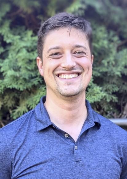 A man in a blue shirt smiling with trees in the background.