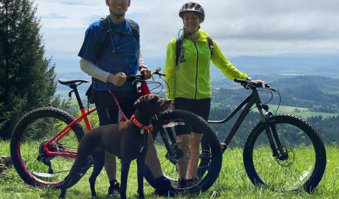 Courtney Rae Armour with husband and dog riding mountain bikes atop grassy hill