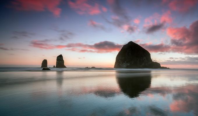 Three rocks jut up from Cannon Beach on the Oregon Coast during a sunset with pink clouds. 