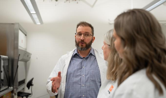 A bearded man wearing glasses and a button-up shirt beneath a lab coat speaks to two other scientists wearing lab coats toward the camera.