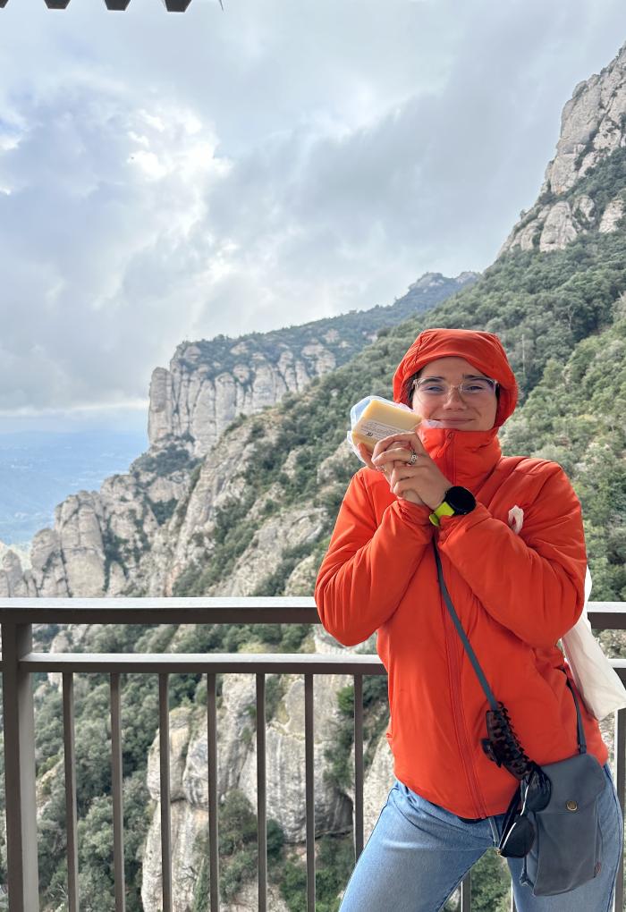A woman in an orange jacket poses for a picture near a mountain holding a package of cheese.