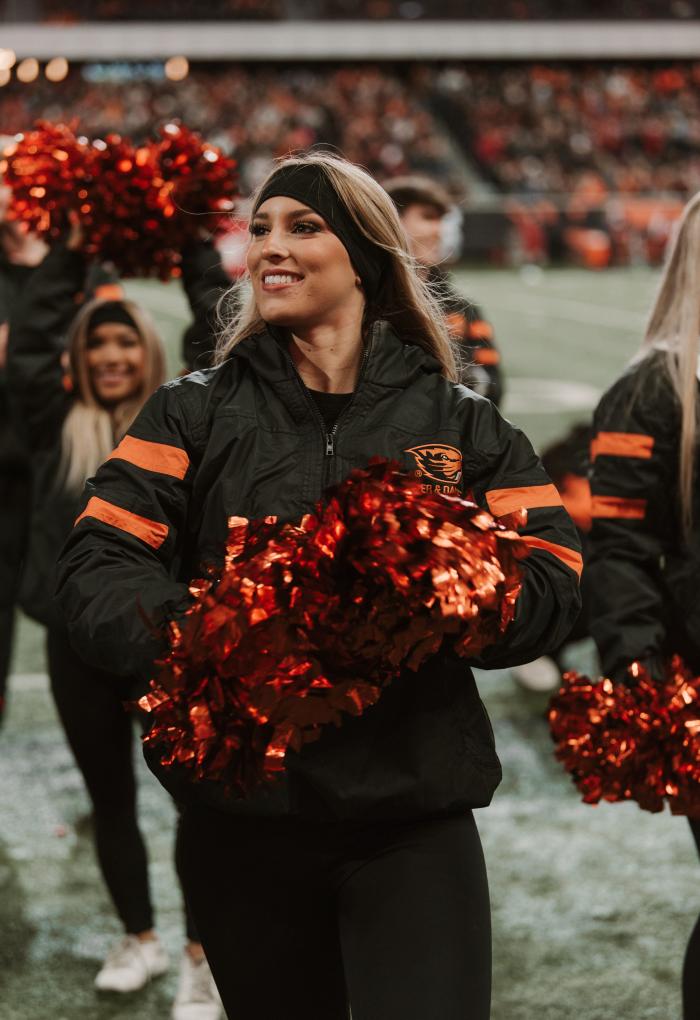 A woman wears a black jacket with orange accents and holds orange-and-black pompoms on a football field, smiling while she looks out to a crowd beyond the camera.