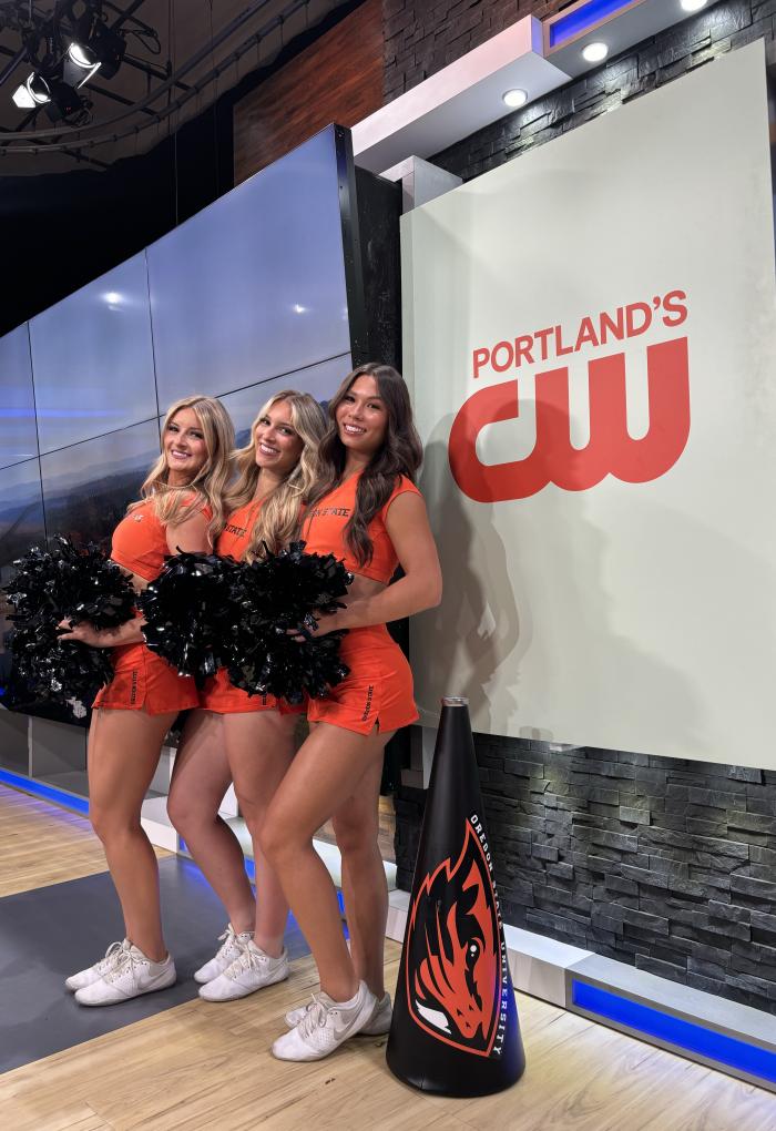 Three woman in orange OSU cheer uniforms stand beside each other, holding pompoms and smiling at the camera in a Portland news studio. 
