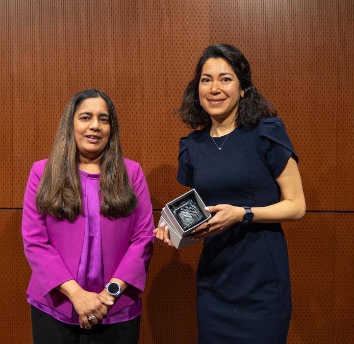 A woman in a purple shirt stands next to a woman in a black outfit holding an award.