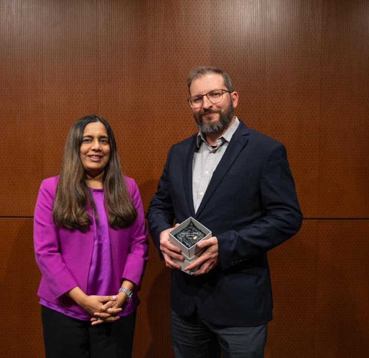 A woman in a purple shirt stands next to a man in a suit holding a glass award.