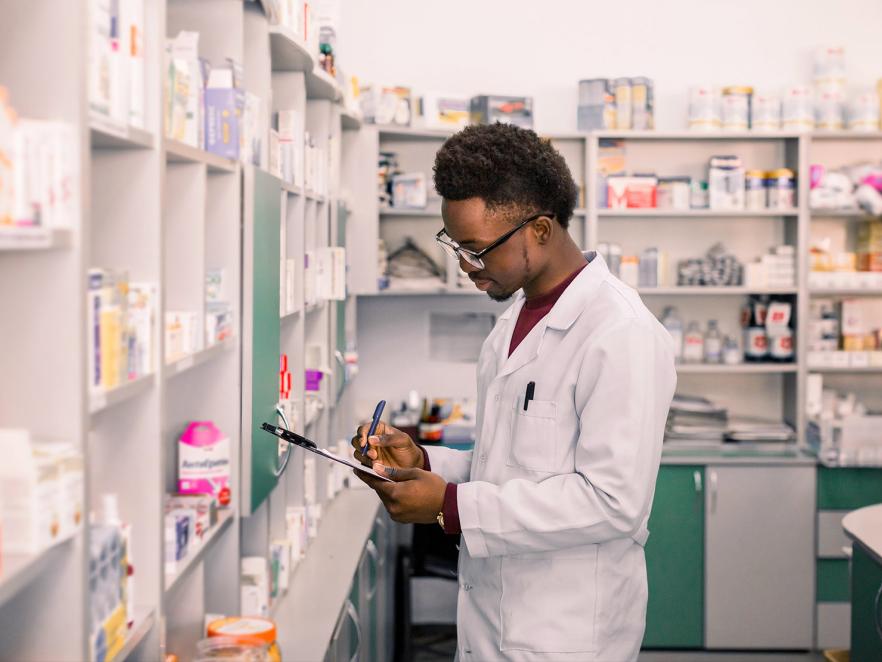 A pharmacist writing on a clipboard next to shelves full of medicine.